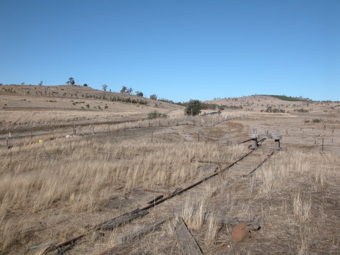 The view looking south from the platform. The formation of the main line is between the siding and the fence. It curves to the left and crosses over the Monaro Highway between the two bushes. The former level crossing here has been removed.