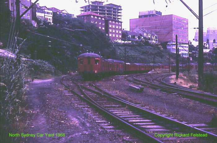 The view looking towards the end of the car sidings, with the near train comprising a mixture of single- and double-deck cars.  The northern pylons of the Harbour Bridge are visible at the far right.